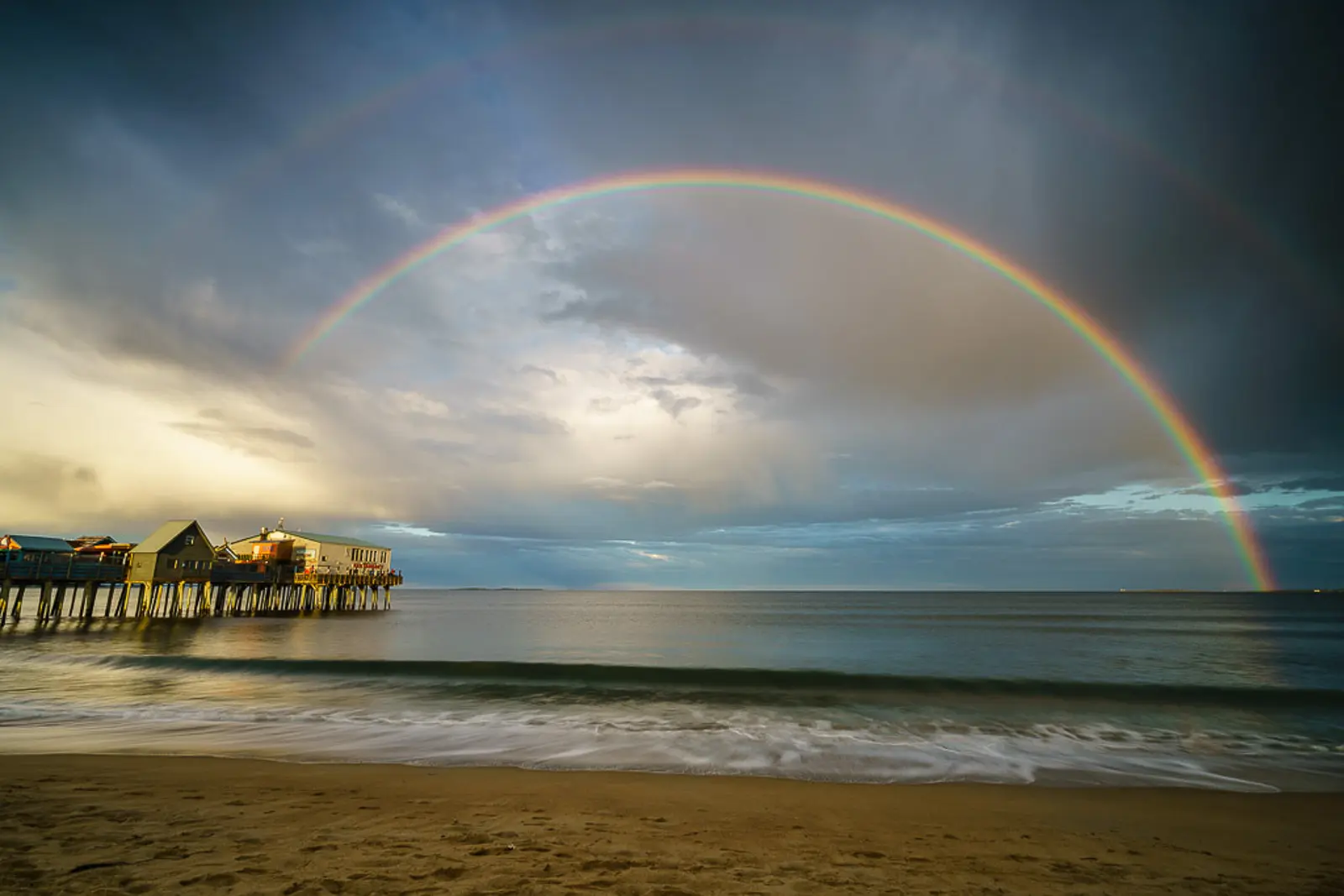 A vibrant double rainbow arcs over Old Orchard Beach, with sunlight reflecting on the sand and water, symbolizing hope and beauty in nature.