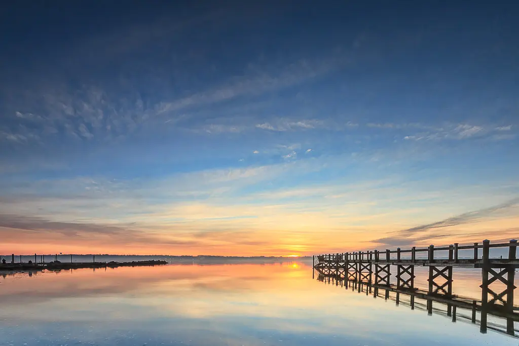 A wooden pier stretching into calm morning water as the sun rises, casting warm golden light across the horizon.