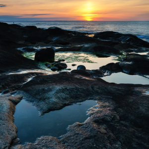 Rocky coastline at sunrise along Narragansett Bay with a heart-shaped tide pool in the rocks and soft golden light through low clouds